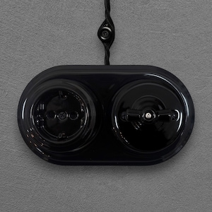 Könnte beinhalten: Eine glänzend schwarze elektrische Wandplatte mit einer kreisförmigen Steckdose auf der linken Seite und einem Drehschalter auf der rechten Seite. Die Einheit ist auf einer strukturierten grauen Oberfläche montiert und hat ein schwarzes Textilkabel.