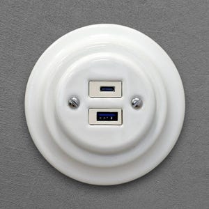 May include: A white, circular USB wall socket with two ports, one USB-C and one USB-A, against a grey wall. The socket has a decorative design with two small screws on either side.