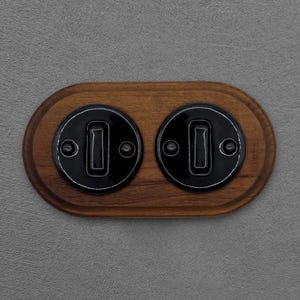 Op de afbeelding: Een houten wandplaat met twee zwarte lichtschakelaars. De schakelaars hebben een rechthoekig ontwerp met een witte omtrek en zijn gemonteerd op een bruine, ovaalvormige houten basis. De achtergrond is een grijze muur.