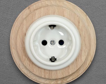 May include: A circular electrical wall socket featuring a glossy white ceramic centre with two pin holes and metal earthing clips, mounted on a light-coloured natural wood base with a stepped edge design and a central silver-coloured screw.