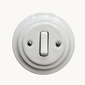 Op de afbeelding: Een ronde witte keramische lichtschakelaar met een glanzende afwerking. De schakelaar heeft een centrale verticale tuimelschakelaar en twee kleine zilverkleurige metalen schroeven aan weerszijden om de afdekplaat aan de basis te bevestigen.