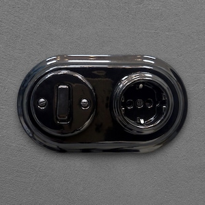 Peut inclure: Un ensemble noir brillant comprenant une prise électrique et un interrupteur, fixé sur un mur gris. La prise comporte trois trous, et l'interrupteur a un bouton rectangulaire. La surface métallique reflète la lumière.
