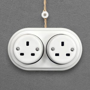 May include: A white, double electrical socket with two circular apertures. The socket is mounted on a grey wall and has a matching white cord and pull. The sockets have three rectangular holes and are secured with small screws.