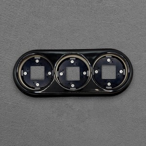 Op de afbeelding: Een glanzend zwart dashboardpaneel met drie ronde openingen, elk voorzien van een vierkante uitsparing in het midden en vier kleine zilverkleurige montageschroeven die symmetrisch in de ronde behuizingsframes zijn geplaatst.