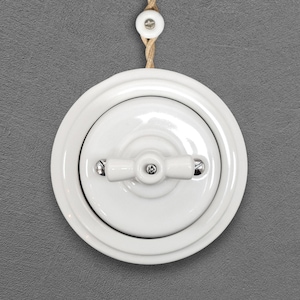 Puede incluir: Un interruptor de luz de cerámica blanca con una palanca de doble brazo, montado en una pared gris. El interruptor es redondo con un borde elevado y está sujeto a un cordón de fibra natural.