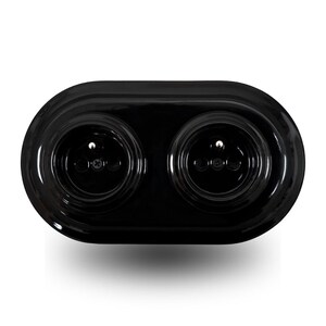 May include: A glossy black double electrical wall socket with an oval-shaped faceplate. The unit features two circular recessed sockets, each containing two pin holes and a central grounding pin, secured by a visible screw in the centre of each.