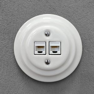Może przedstawiać: Biała, okrągła, ceramiczna płyta ścienna z dwoma portami Ethernet RJ45. Płyta jest zamontowana na szarej, teksturowanej ścianie i zabezpieczona dwoma srebrnymi śrubami. Porty mają beżowe wnętrze.