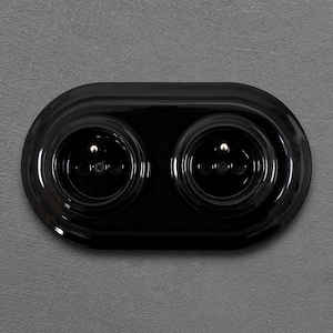 Peut inclure: Une prise électrique double noire brillante avec deux prises circulaires. La prise est encastrée dans une plaque ovale et montée sur un mur gris. Les prises ont de petits détails de vis.
