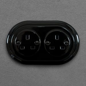 May include: A black double electrical socket with two circular apertures. The socket is set in an oval plate, mounted on a grey textured wall. Each socket has three rectangular holes for plugs.