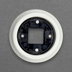 Pode incluir: Uma tampa de tomada elétrica branca e circular com interior preto. A tampa tem uma abertura quadrada no centro e é fixada com quatro parafusos prateados. O item está sobre um fundo cinza.