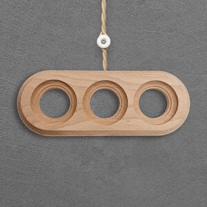 Op de afbeelding: Een lichtbruine houten houder met een langwerpige ovale vorm en drie ronde uitsparingen. De houder hangt aan een gedraaid beige koord met een kleine witte ronde sluiting met een zichtbare schroef in het midden.