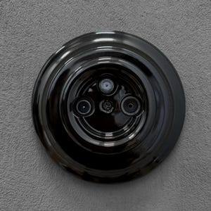 Könnte beinhalten: Eine glänzend schwarze Steckdose mit drei kreisförmigen Öffnungen und einer zentralen Schraube. Die Steckdose ist an einer strukturierten grauen Wand montiert und zeigt eine Vintage- oder Industrie-Designästhetik.
