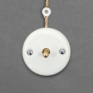 Peut inclure: Un interrupteur de lumière rond blanc avec une bascule en laiton et deux vis argentées. Une pièce en céramique blanche assortie est fixée à une corde beige au-dessus de l'interrupteur. L'interrupteur est posé sur un fond gris.