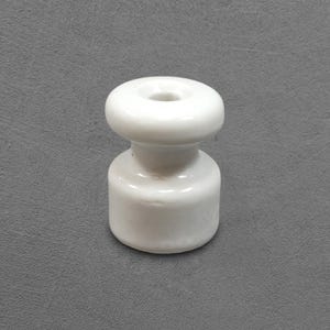 Peut inclure: Un isolateur en céramique blanc, avec une base cylindrique et un dessus arrondi percé d'un trou central. L'objet est lisse et brillant, posé sur un fond gris. Il est probablement utilisé dans les applications électriques.