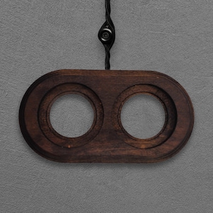 Pode incluir: Um suporte de madeira castanho-escuro com formato oval alongado e dois recortes circulares. A peça está suspensa por um cordão trançado preto que se prende a um pequeno componente circular preto centrado acima da moldura de madeira.