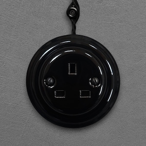 May include: A black electrical socket with a glossy finish, featuring three rectangular slots and two screw details. The socket is mounted on a grey wall and connected to a black cord with a button.