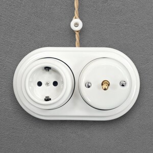 May include: A white ceramic double wall plate featuring a circular electrical socket on the left and a toggle light switch with a brass lever on the right. The unit is mounted on a grey surface and includes a braided tan cord with a white ceramic bead.