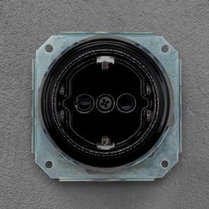 Peut inclure: Gros plan d'une prise électrique noire avec trois trous circulaires et une vis centrale. La prise est encastrée dans un cadre métallique carré avec des coins arrondis et des trous de montage. Le fond est une surface grise texturée.