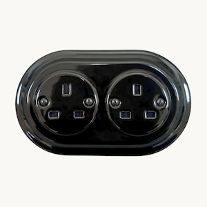 May include: A glossy black double electrical wall socket with an oval-shaped faceplate. The unit features two standard three-pin socket configurations, each secured with two visible screws, presenting a smooth, reflective surface finish.