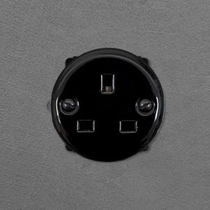 Op de afbeelding: Een cirkelvormig zwart stopcontact met een standaard configuratie van drie gaten. De glanzende, ronde afdekplaat is bevestigd met twee zichtbare zwarte schroeven aan de linker- en rechterkant, gecentreerd op het gladde, donkere oppervlak.
