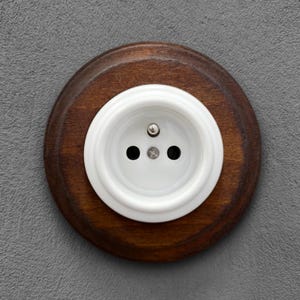 Peut inclure: Une prise électrique blanche à trois trous est encastrée dans une base en bois circulaire brun foncé. La prise est montée sur un mur gris texturé, créant un contraste.