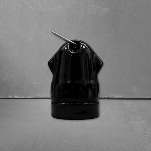 Op de afbeelding: Een glanzend zwart keramisch object met een afgeronde bovenkant en een cilindrische basis. Een dunne, zilverkleurige metalen pin is in het midden van de bovenkant gestoken. Het stuk heeft twee kleine, symmetrische uitsteeksels aan de zijkanten.
