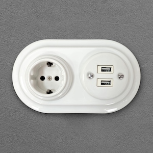 Könnte beinhalten: Eine weiße, ovale Wandsteckdose mit einer runden Standard-Steckdose auf der linken Seite und zwei übereinander angeordneten USB-Anschlüssen auf der rechten Seite. Die Einheit ist mit zwei sichtbaren Metallschrauben rechts und einer mittig befestigt.