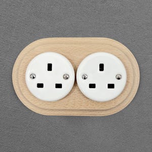 Peut inclure: Une double prise électrique avec des plaques frontales blanches et des fentes noires est encastrée dans une base ovale en bois clair. Les prises ont des vis argentées. Le design est moderne et minimaliste.