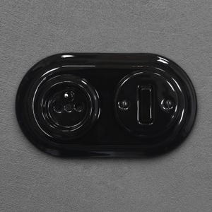 Peut inclure: Une prise électrique et un interrupteur noirs brillants. La prise comporte trois trous circulaires, et l'interrupteur est un rectangle vertical. La plaque est de forme ovale et montée sur une surface grise texturée.