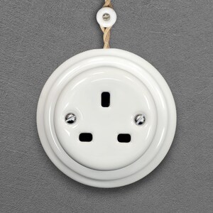 May include: A round, white ceramic electrical wall socket featuring three rectangular pin slots and two silver-coloured metal screws. The socket is mounted on a textured grey surface and is connected to a braided tan cord with a small white bead.