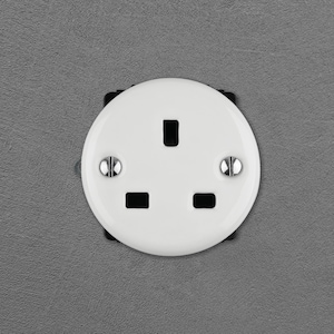 Może przedstawiać: Okrągłe, białe gniazdko elektryczne ścienne z trzema otworami ułożonymi w trójkąt. Płyta czołowa jest przymocowana za pomocą dwóch płaskich metalowych śrub umieszczonych po bokach i posiada gładkie, błyszczące białe wykończenie.