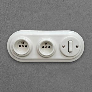 Op de afbeelding: Een witte, ovaalvormige wandplaat met twee stopcontacten en één tuimelschakelaar. De unit is afgewerkt met een gladde, glanzende laag en is bevestigd met zichtbare metalen schroeven op het schakelaarcomponent.