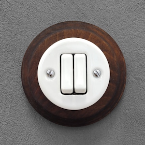 Op de afbeelding: Een lichtschakelaar in vintage stijl met twee witte tuimelschakelaars in een witte plaat. De plaat is gemonteerd op een ronde, donkerbruine houten basis. Zilveren schroeven bevestigen de plaat aan het hout. De achtergrond is een grijze muur.