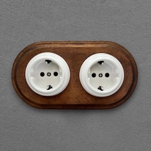 May include: A wooden electrical outlet featuring two white sockets. The sockets are set in a dark brown, oval-shaped wooden plate against a grey wall. The sockets have three holes for plugs.