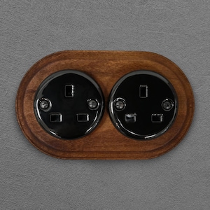 May include: A double electrical socket with black faceplates set in a dark wood oval plate. The sockets are glossy and mounted on a grey wall. The wood has a natural grain and is stained.