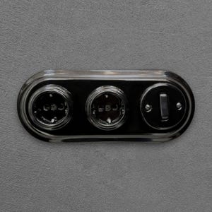 Op de afbeelding: Een glanzend zwarte elektrische wandplaat met twee ronde stopcontacten en één tuimelschakelaar. De unit is horizontaal gemonteerd op een grijs gestructureerd oppervlak, met zichtbare schroeven die de schakelaar en de stopcontacten bevestigen.
