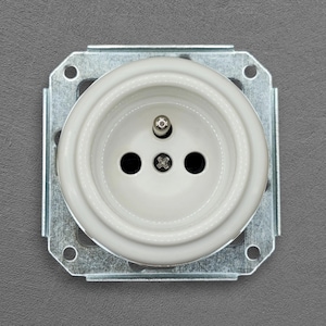 Peut inclure: Une prise électrique blanche avec trois trous et une vis centrale, montée sur une plaque métallique carrée. La prise est conçue pour les fiches électriques. L'image est une prise de vue rapprochée.