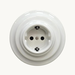 Puede incluir: Un enchufe eléctrico de pared circular de cerámica blanca con un diseño escalonado. El centro presenta dos orificios redondos y dos clips de conexión a tierra de metal negro colocados arriba y abajo, asegurados por un tornillo central plateado.