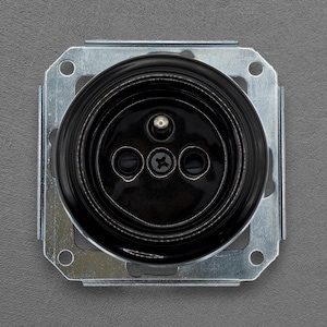 May include: A black ceramic light socket with three screw holes, set within a square metal mounting plate. The socket has a glossy finish and is mounted on a grey surface. The design is simple and functional.