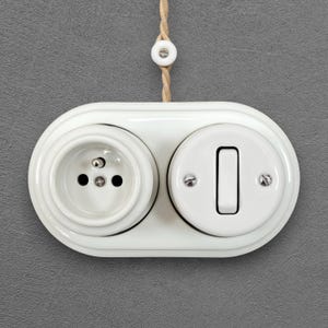 Può includere: Una placca a muro doppia in ceramica bianca con una presa elettrica circolare a sinistra e un interruttore a levetta rettangolare a destra. L'unità è montata su una superficie grigia e include un cavo in tessuto intrecciato color beige con perla.