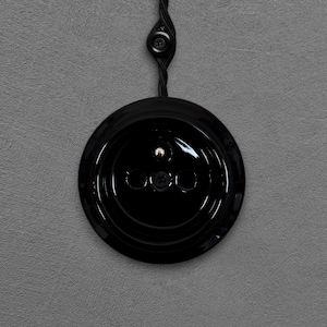 Peut inclure: Un interrupteur de lumière noir et rond, avec une finition brillante, comportant trois trous de vis et un petit détail métallique. L'interrupteur est relié à un cordon noir torsadé, sur un mur gris texturé.