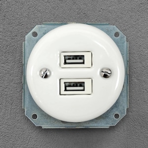 May include: A white circular wall-mounted electrical socket featuring two stacked USB ports. The faceplate is secured by two silver-coloured metal screws, and the entire unit is mounted on a square metallic frame with visible mounting holes.