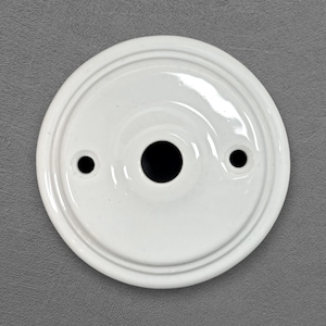 Pode incluir: Uma placa de parede circular de cerâmica branca com acabamento brilhante, uma abertura central e dois orifícios de montagem menores posicionados em lados opostos. A placa apresenta um design de anéis concêntricos escalonados.