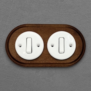 Op de afbeelding: Een ovale houten wandplaat met twee witte, ronde lichtschakelaars. Elke schakelaar heeft een witte tuimelschakelaar en is bevestigd met kleine zilveren schroeven. Het hout is donkerbruin gebeitst.