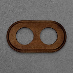Könnte beinhalten: Eine ovale Steckdosenabdeckung aus Holz mit zwei kreisförmigen Öffnungen. Die Abdeckung hat eine warme braune Farbe und einen leicht erhöhten, abgerundeten Rand. Der Hintergrund ist eine strukturierte graue Oberfläche.