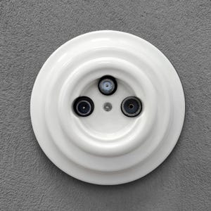 Peut inclure: Une prise électrique blanche et circulaire avec trois ports encastrés. La prise est montée sur un mur gris texturé. Les ports sont noirs et argentés, et la prise a un design à plusieurs niveaux.
