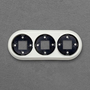 Può includere: Una copertura bianca per presa elettrica con tre aperture circolari, ciascuna con un centro quadrato nero. La copertura è rettangolare con estremità arrotondate, su sfondo grigio. Design moderno e minimalista.