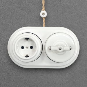 Op de afbeelding: Een witte dubbele wandplaat van keramiek met een rond stopcontact aan de linkerkant en een draaischakelaar aan de rechterkant. De unit is gemonteerd op een grijs gestructureerd oppervlak en bevat een beige gevlochten snoer met een keramische kraal.