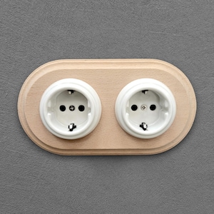 Peut inclure: Une prise électrique double avec des prises en céramique blanche encastrées dans une plaque ovale en bois clair. Les prises ont trois trous et des broches métalliques. La plaque est fixée sur un mur gris.