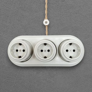 Könnte beinhalten: Eine weiße Dreifachsteckdose mit drei runden Steckdosen. Die Steckdose ist an einer grauen Wand befestigt und mit einem beigefarbenen Kabel verbunden. Die Steckdosen haben zwei Löcher für Stecker.